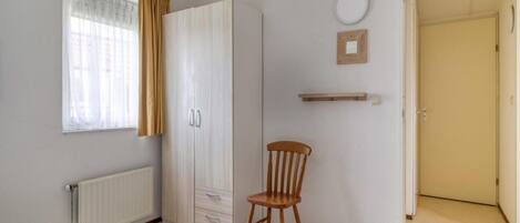 Un dormitorio con cama, armario, silla, radiador, ventana con cortinas, puerta y estante fijado a la pared.