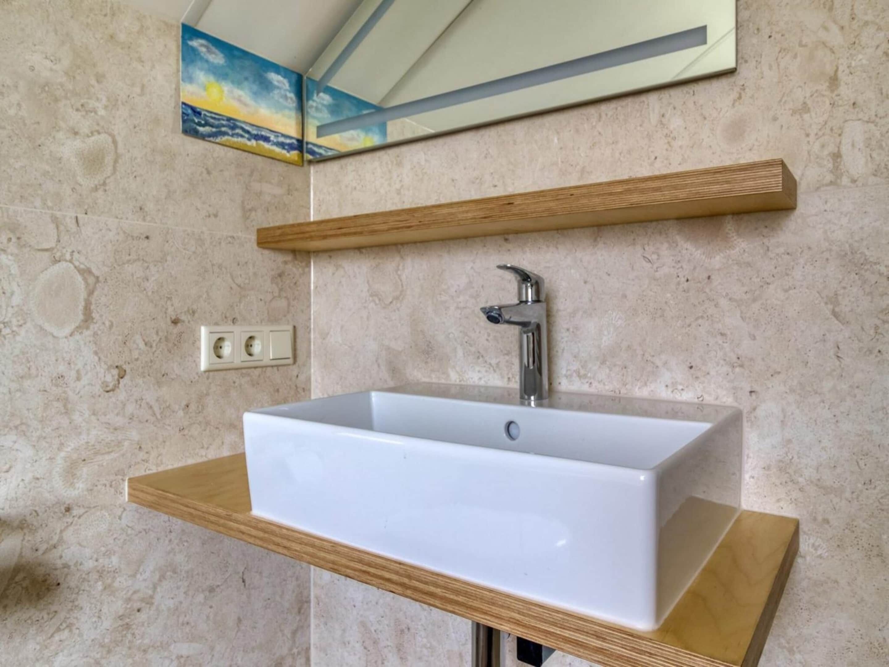 Une salle de bain avec un lavabo rectangulaire blanc, une étagère en bois au-dessus et un miroir fixé au mur.