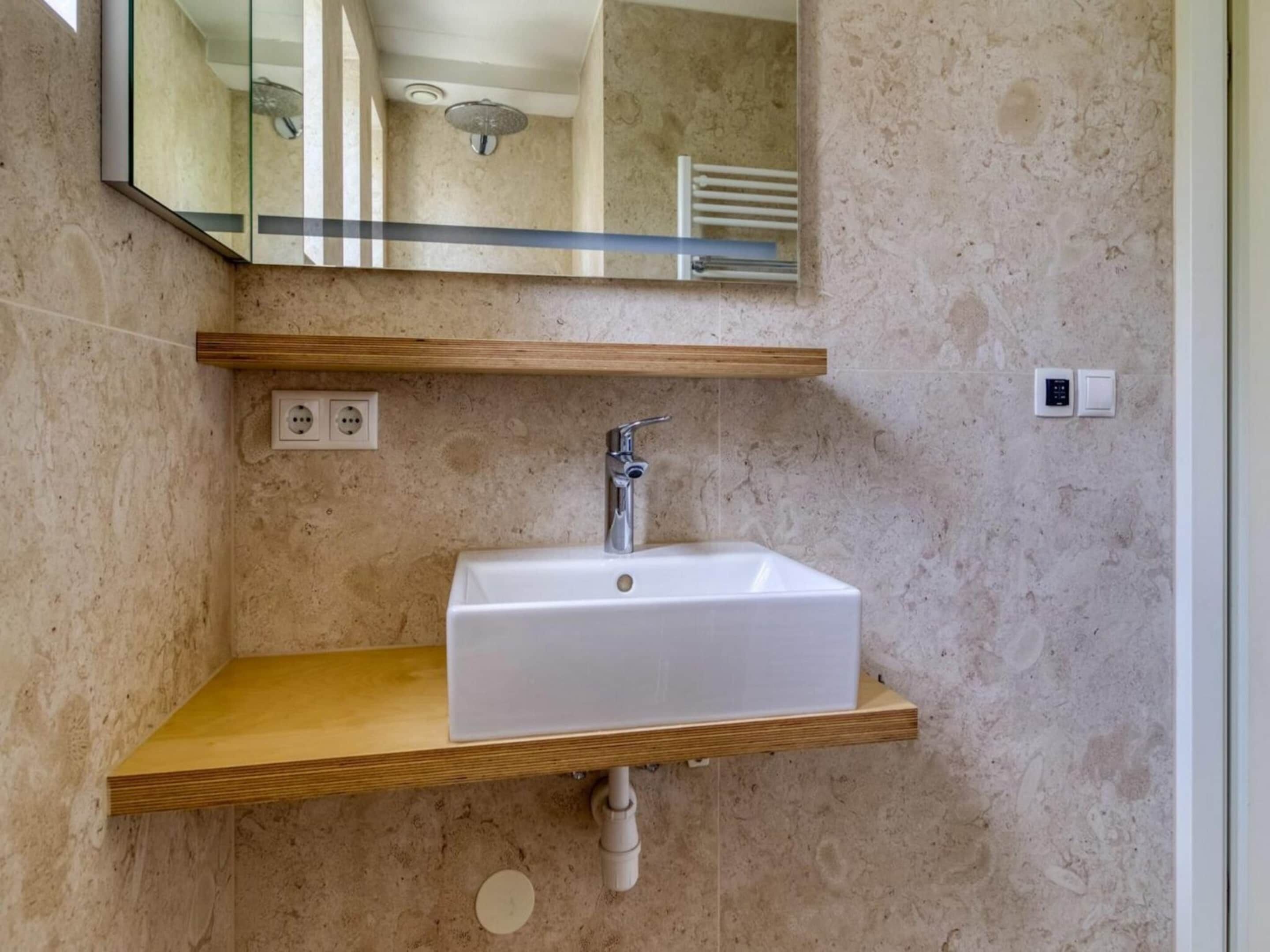 A bathroom with a white sink, wooden shelf, and a mirror reflecting a shower area.