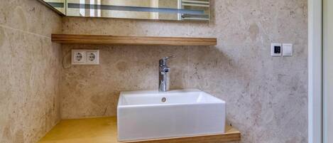 Une salle de bain avec un lavabo blanc, une étagère en bois et un miroir qui reflète l’espace de la douche.