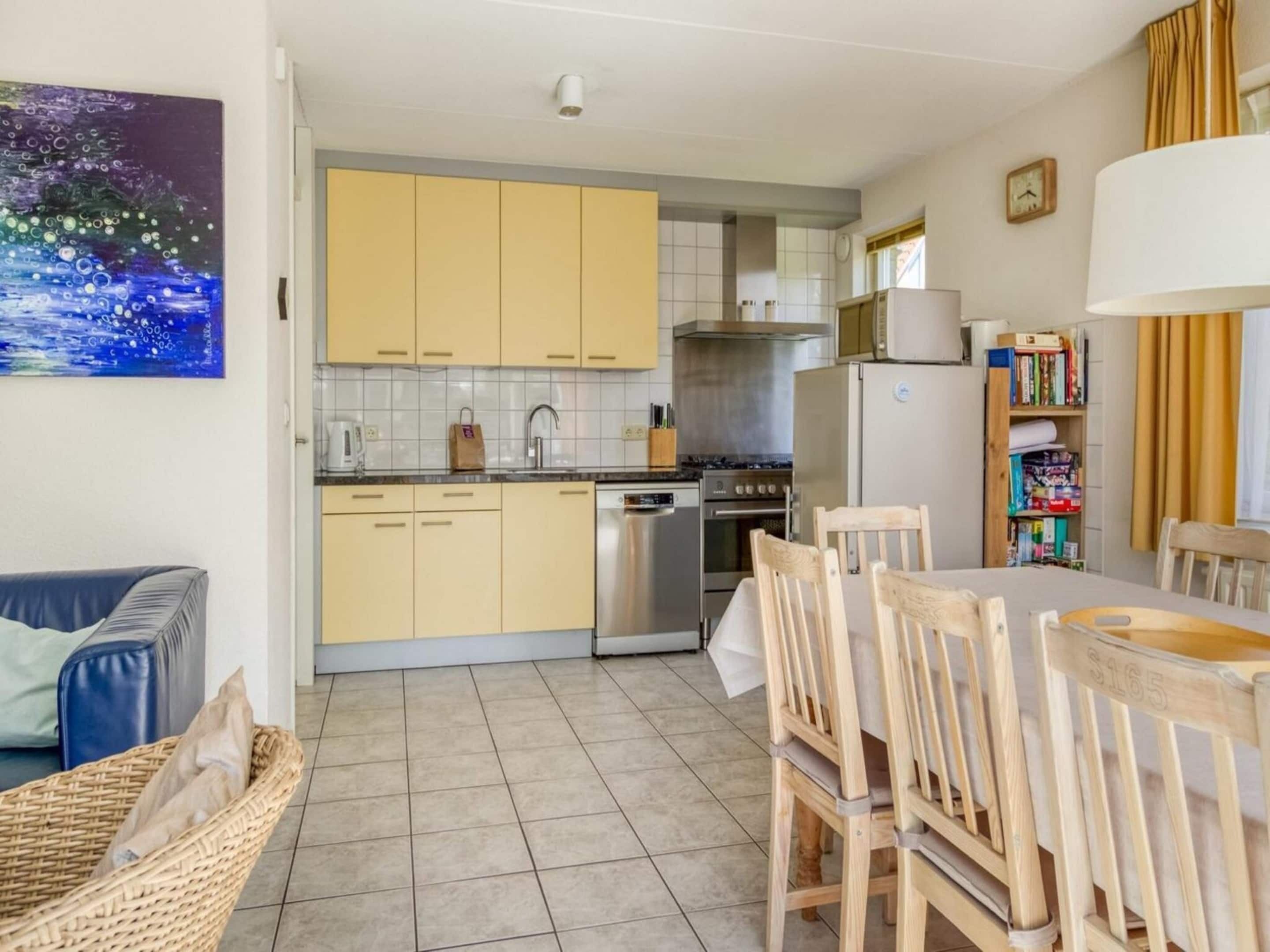 A compact kitchen with modern appliances, a dining area with wooden chairs, and a bookshelf.