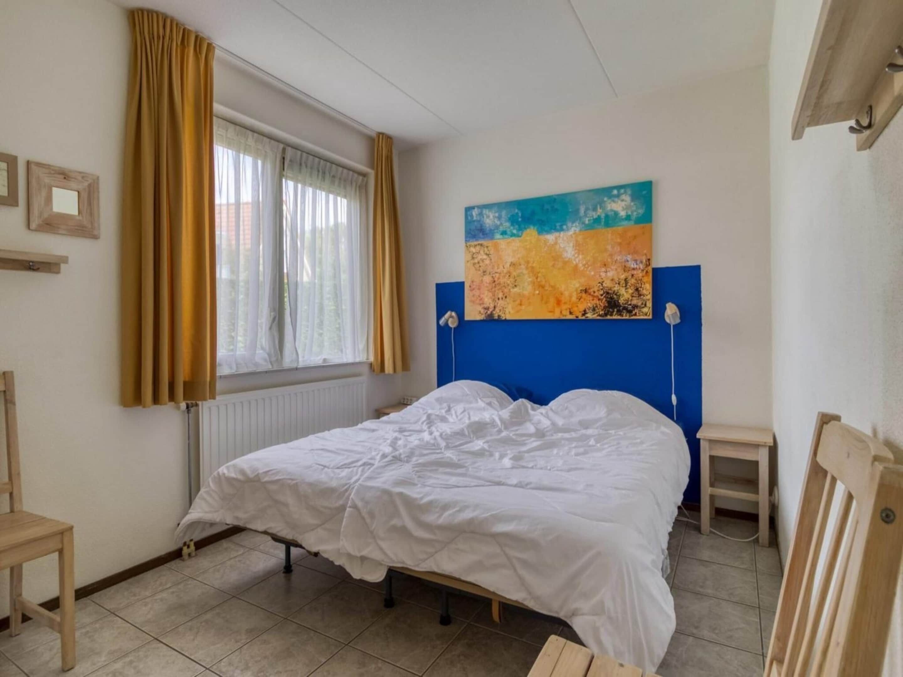 A bedroom with a bed, a window with curtains, a blue headboard, and a painting on the wall.