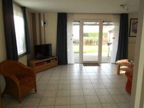 A living room with a television, a wicker chair, a small table, and a sliding glass door leading to an outdoor area. - Lovely Bungalow With Garden (Breskens)