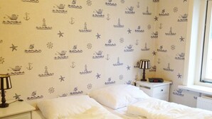 A bedroom with a bed, white bedding, a nightstand with a lamp, and a wall with nautical-themed wallpaper. - Historic Cottage w/ Garden (Breskens)