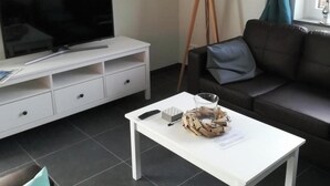 A modern living room with a flat-screen TV, white furniture, a standing lamp, a sofa, and a coffee table with items on it. - Bungalow in Breskens With Private Jetty (Breskens)