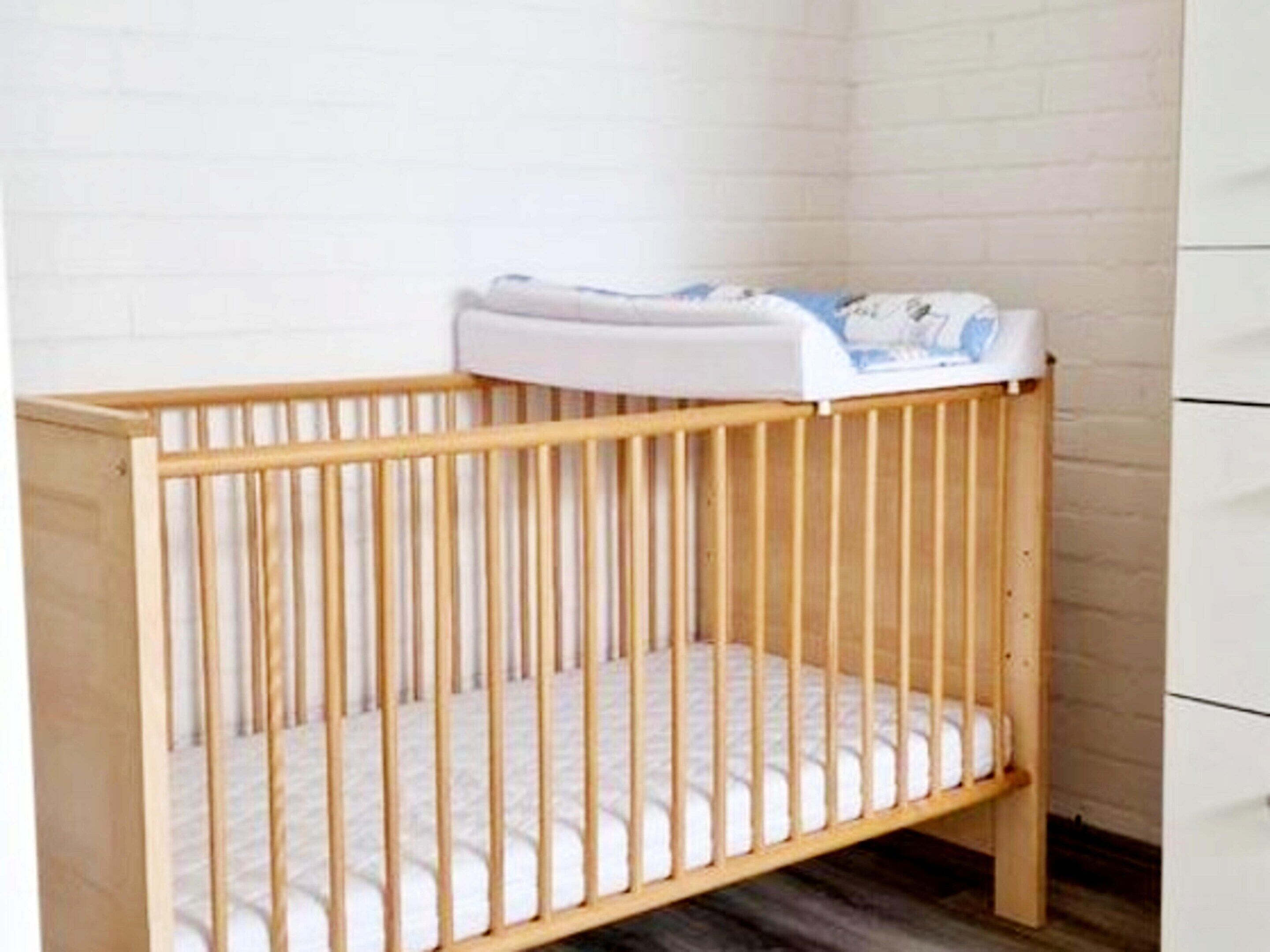 A wooden crib with a white mattress and a white headboard against a white brick wall.