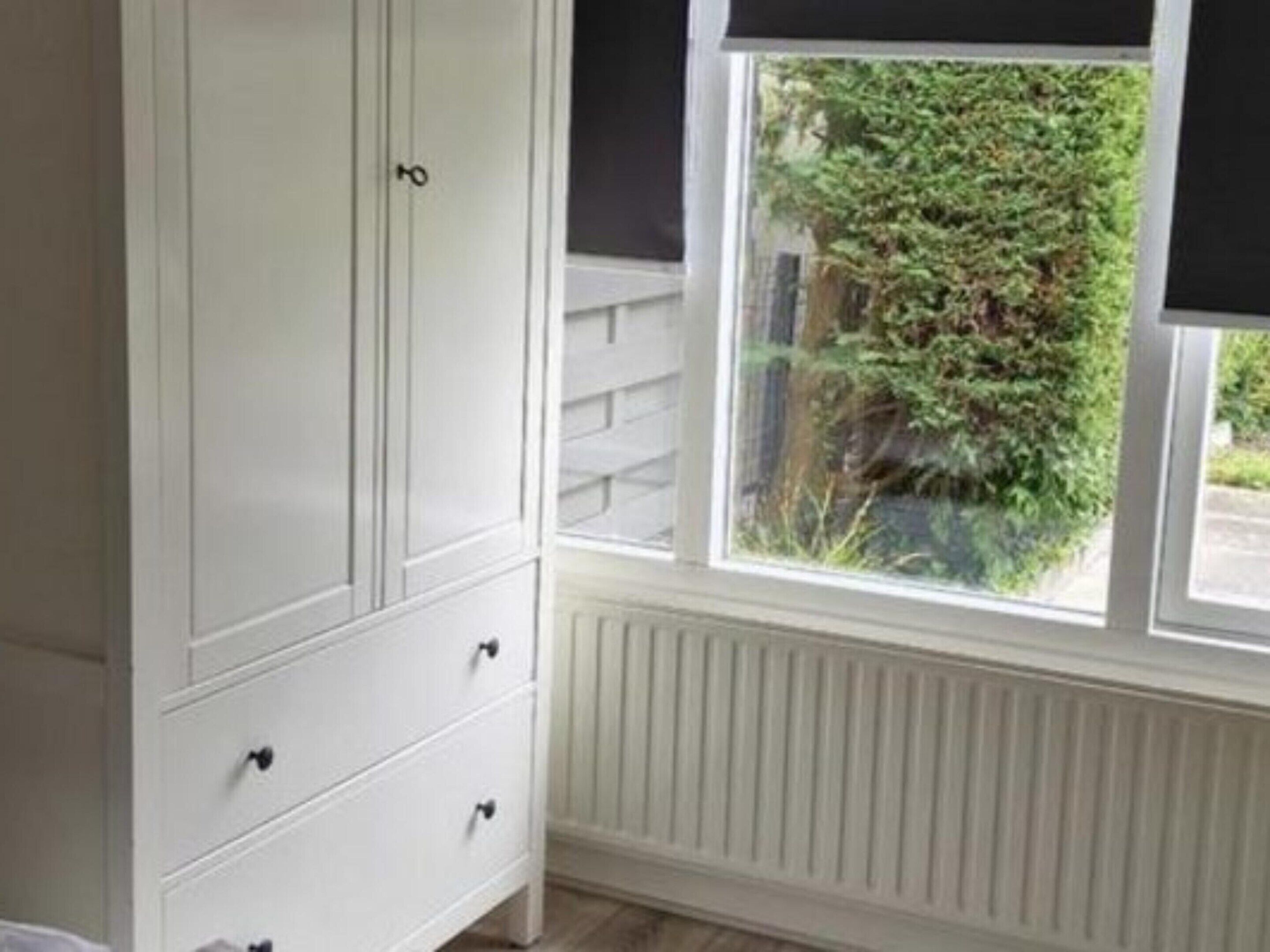 A room with a white wardrobe, a window with a view of greenery, and a radiator.