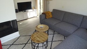 A living room with a grey sofa, a TV on a white cabinet, two round wooden tables, a fireplace, and a window with curtains. - Family Stay - Near Beach (Breskens)