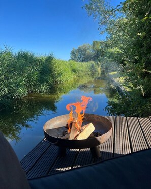 A fire pit on a wooden deck by a calm river surrounded by lush greenery. - Holiday Home on Waterfront in Zeeland (Groede)