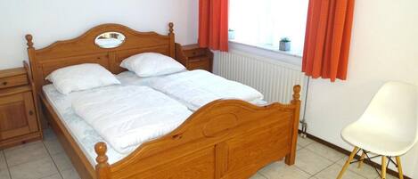 Una cama con dosel de madera, ropa de cama blanca, una mesita de noche, una silla blanca y una ventana con cortinas rojas.