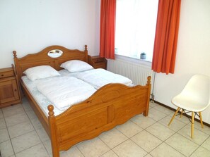 A wooden four-poster bed with white bedding, a nightstand, a white chair, and a window with red curtains. - Bungalow in Breskens Near Beach and Lighthouse (Breskens)
