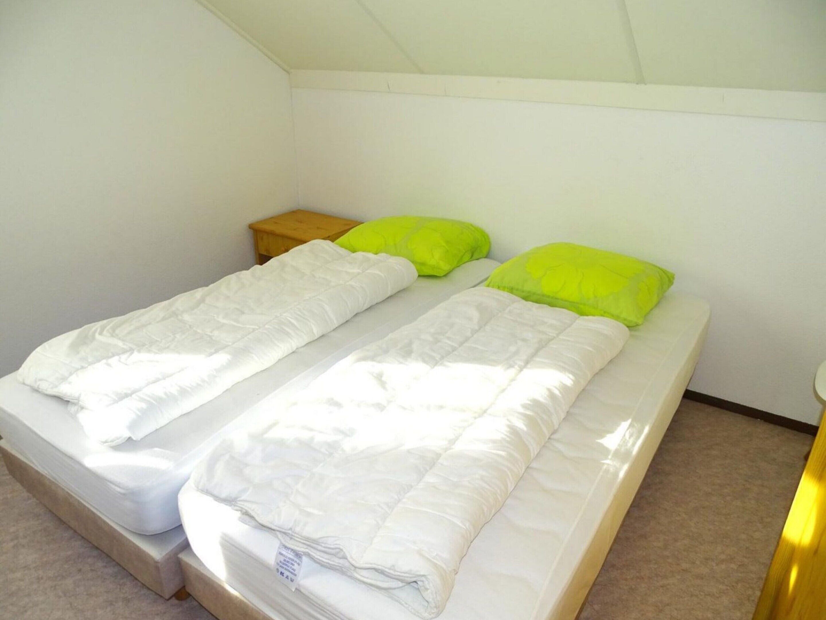 Una cama doble con ropa de cama blanca y dos almohadas verde brillante en una habitación pequeña y sencilla con paredes blancas y una mesita de noche de madera.