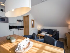 A modern kitchen with white cabinets, a microwave, and a stove. There is a dining area with a wooden table and chairs, and a sofa in the background.