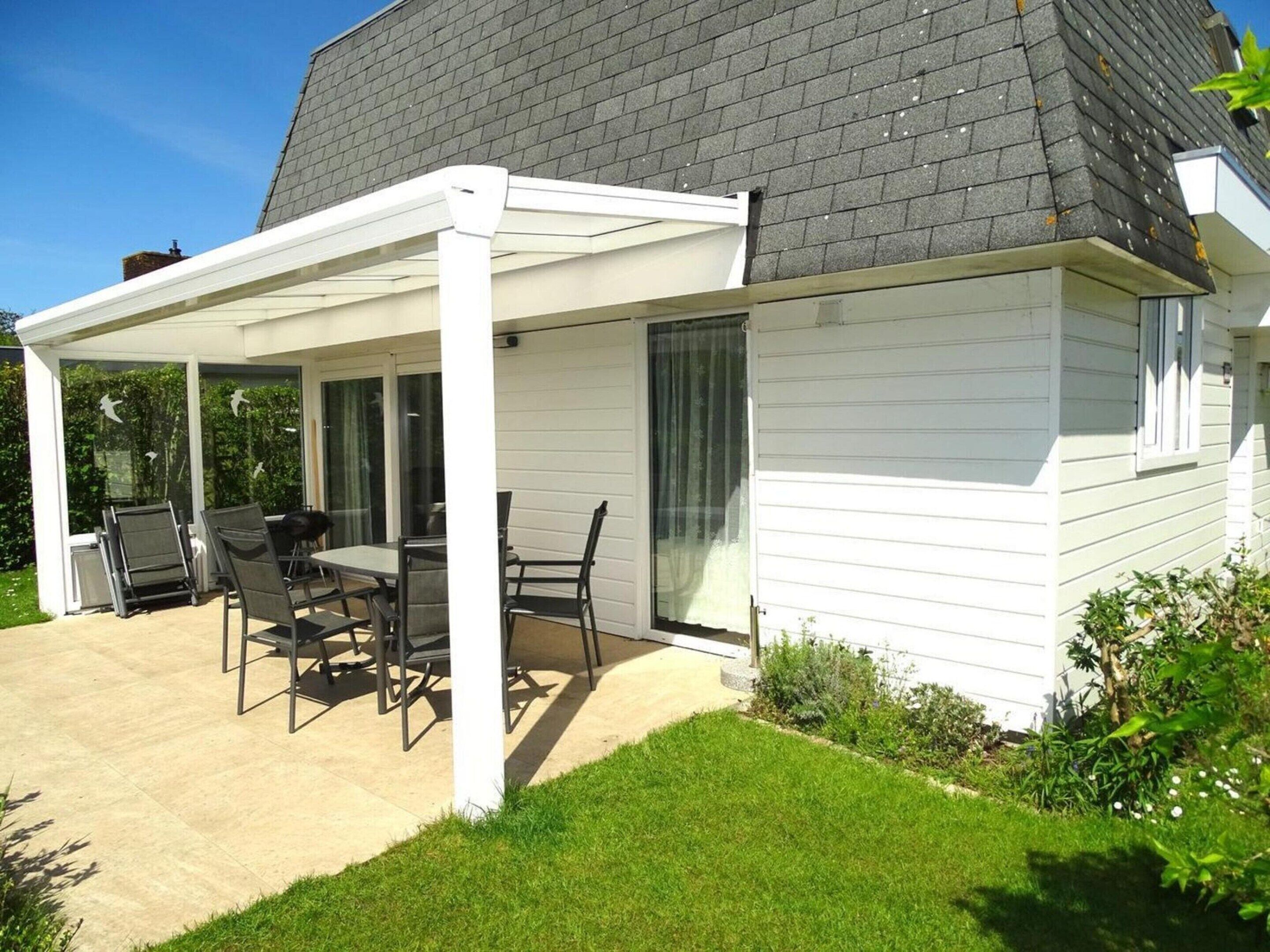 A covered patio with a table and chairs, a white exterior wall, and a dark roof.