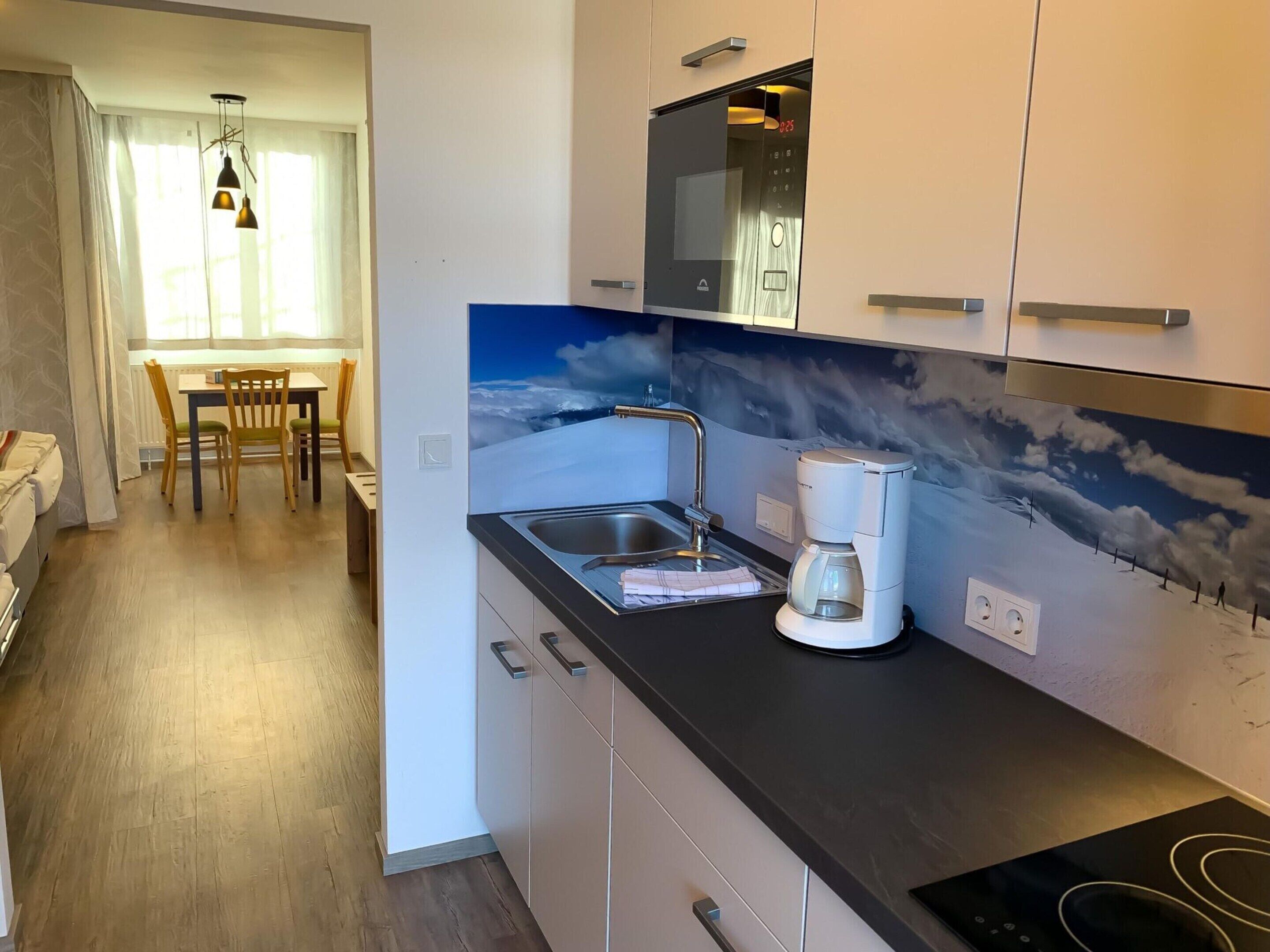 Una cocina moderna con microondas integrado, cafetera y un lavabo. Se ve un comedor con una mesa y sillas al fondo.
