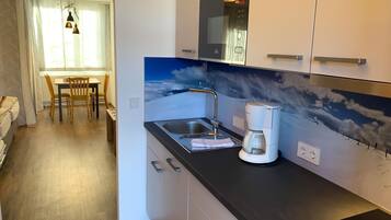 Una cocina moderna con microondas integrado, cafetera y un lavabo. Se ve un comedor con una mesa y sillas al fondo.
