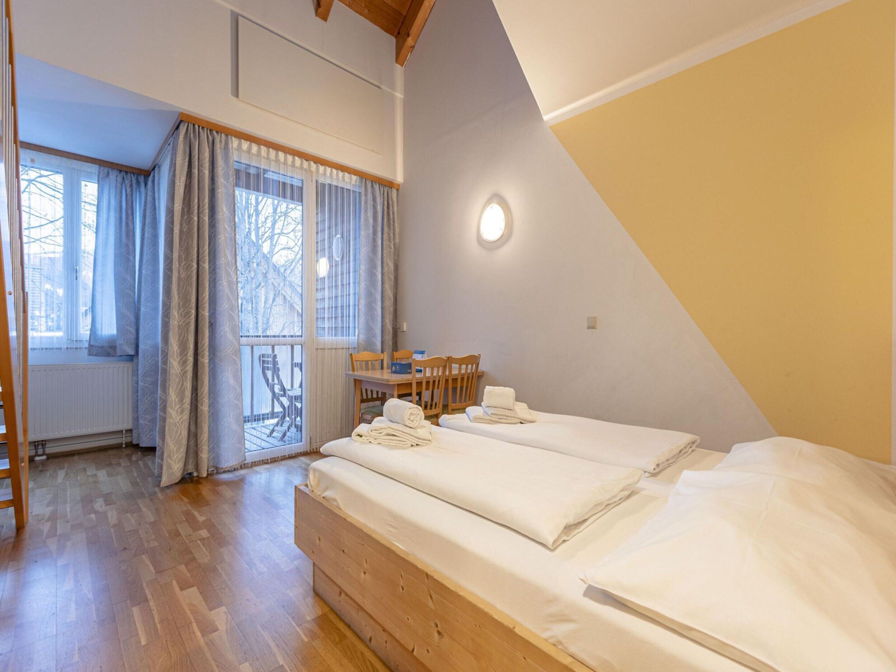 Ein Schlafzimmer mit zwei Betten, Holzboden, Essecke mit Stühlen und Blick auf Bäume durch das Fenster.