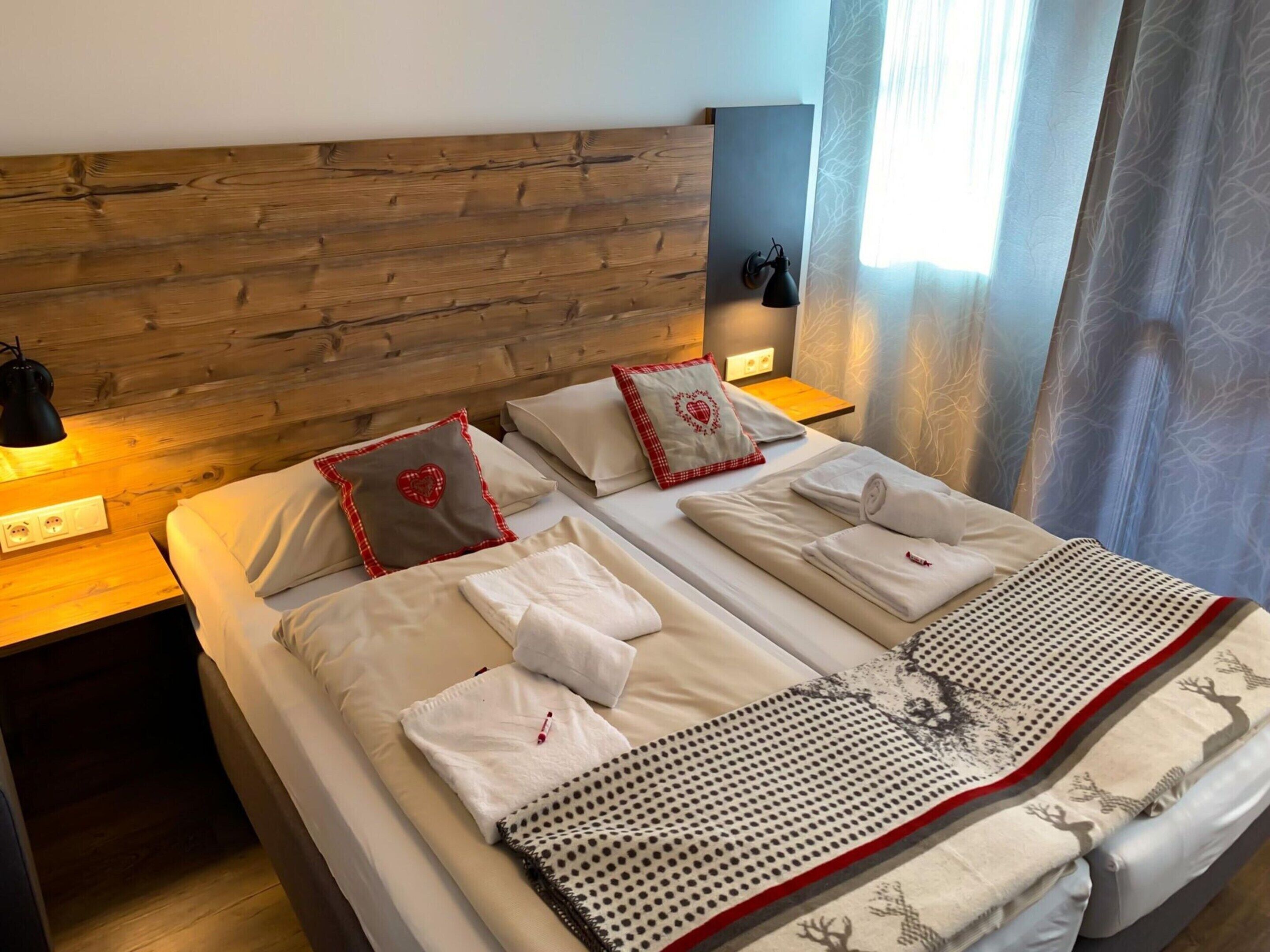 Una cama doble con sábanas blancas, dos almohadas con diseños de corazones, un edredón con estampado de renos, una lámpara de pared y una ventana con cortinas traslúcidas.