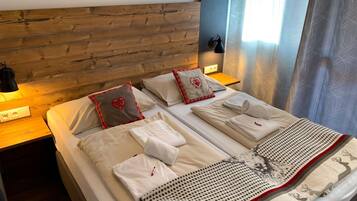 Una cama doble con sábanas blancas, dos almohadas con diseños de corazones, un edredón con estampado de renos, una lámpara de pared y una ventana con cortinas traslúcidas.