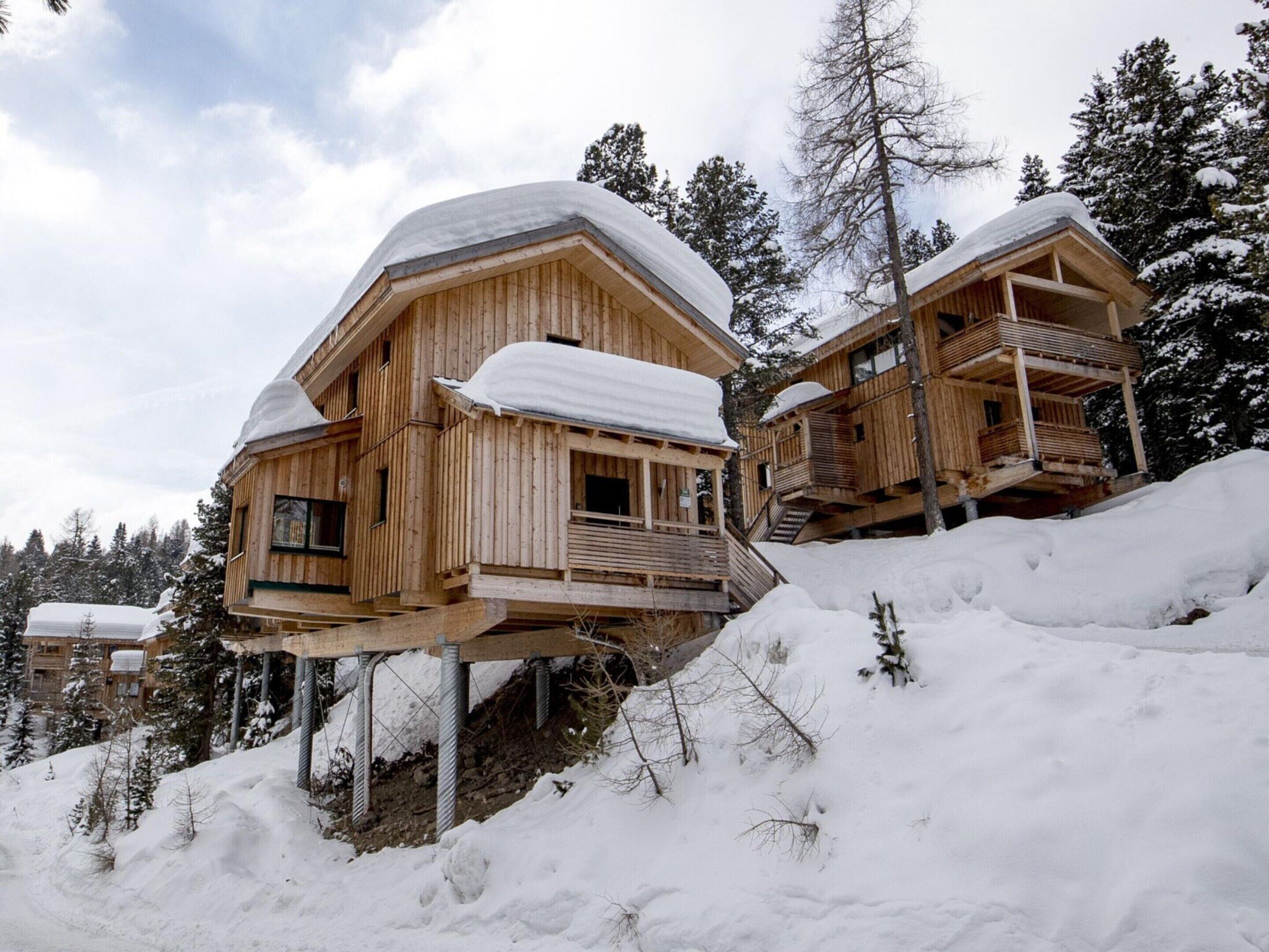 Two wooden cabins with snow-covered roofs, elevated on stilts, surrounded by snow and trees.