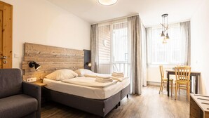 Een moderne hotelkamer met een bed met houten hoofdeinde, een grijze bank, een houten eettafel met stoelen, en een groot raam met gordijnen.