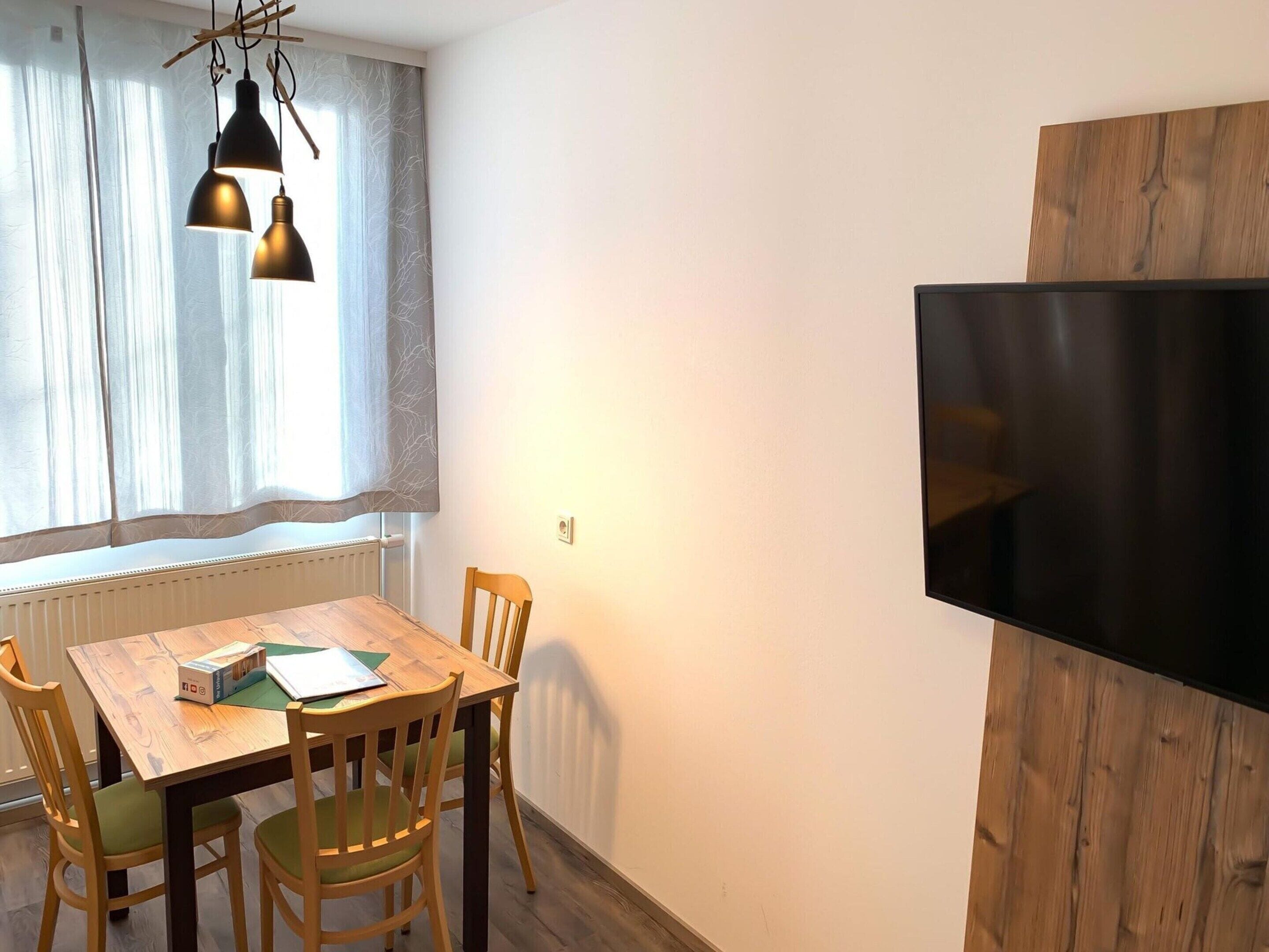 Un comedor con una mesa y sillas de madera, una ventana con cortinas ligeras y un televisor de pantalla plana montado en la pared.