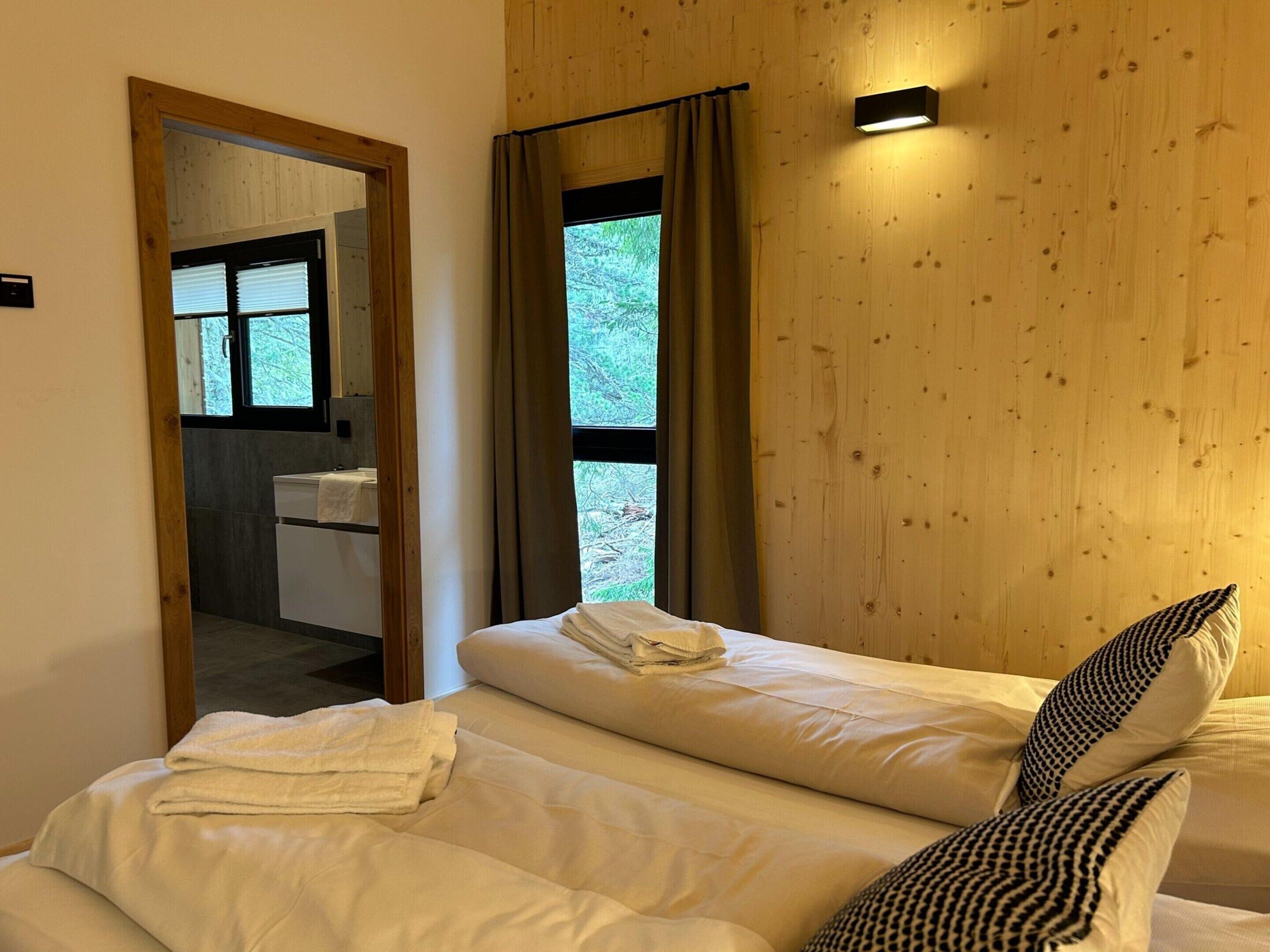 A cozy bedroom with a wooden wall, a large window with curtains, a bedside table, and a mirror reflecting a bathroom with a sink.
