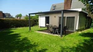 Une terrasse couverte avec une table et des chaises, un petit bâtiment doté d’une porte et de fenêtres, et une cour gazonnée.