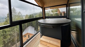 A modern hot tub on a balcony with large windows overlooking a forested mountain area.