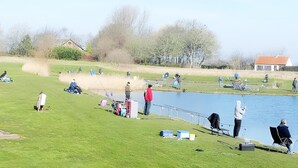 Ein See mit einem Angelplatz, Personen beim Angeln, einem kleinen Haus und einem grasbewachsen Feld.