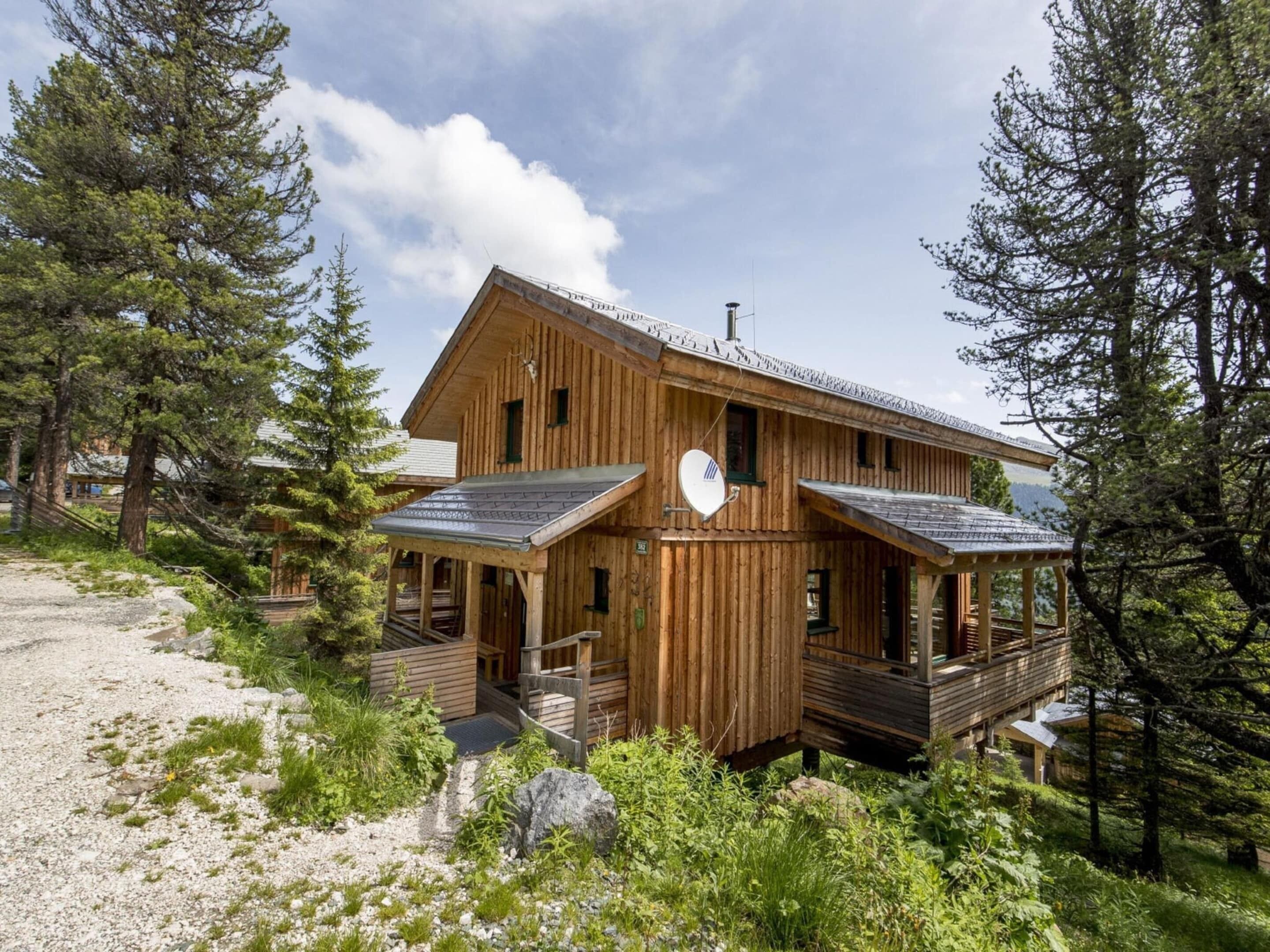 A wooden cabin with a deck, surrounded by trees and a gravel path.