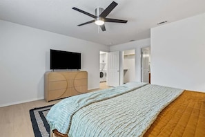 Un dormitorio con cama, ventilador de techo, televisor, armario y lavadora al fondo.