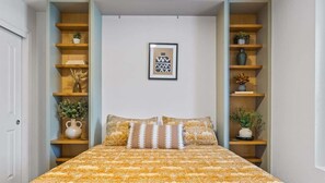 A bedroom with a bed, a wall-mounted shelf with decorative items, and a framed picture above the bed.