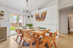 A dining area with a wooden table set for six, surrounded by chairs. There is a large window with a view of a backyard.
