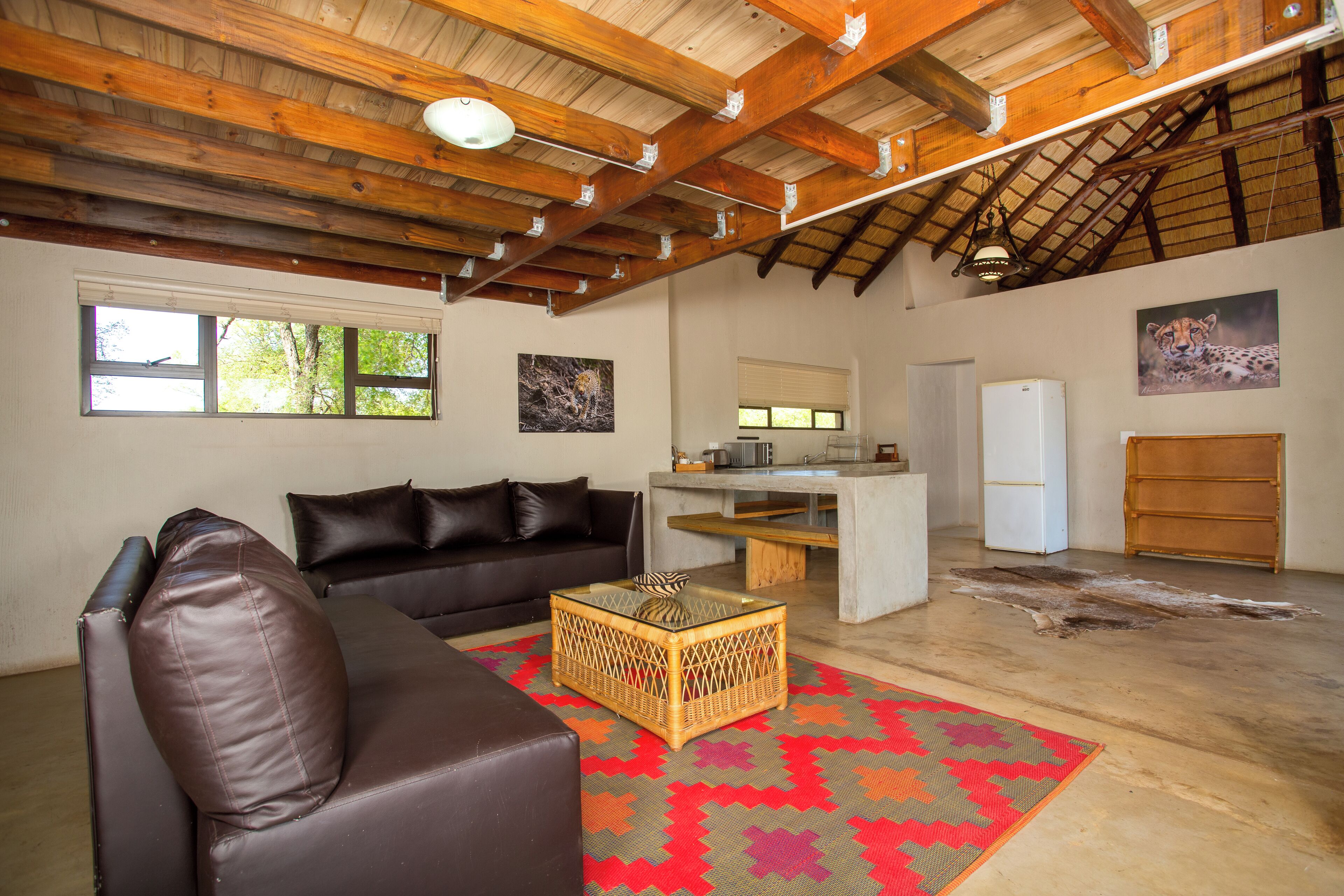 A spacious living room with a brown leather sofa, a wicker coffee table, and a colorful rug. There is a kitchen area with a refrigerator and a wooden cabinet.