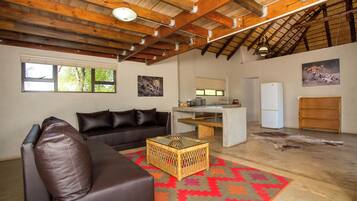 A spacious living room with a brown leather sofa, a wicker coffee table, and a colorful rug. There is a kitchen area with a refrigerator and a wooden cabinet.