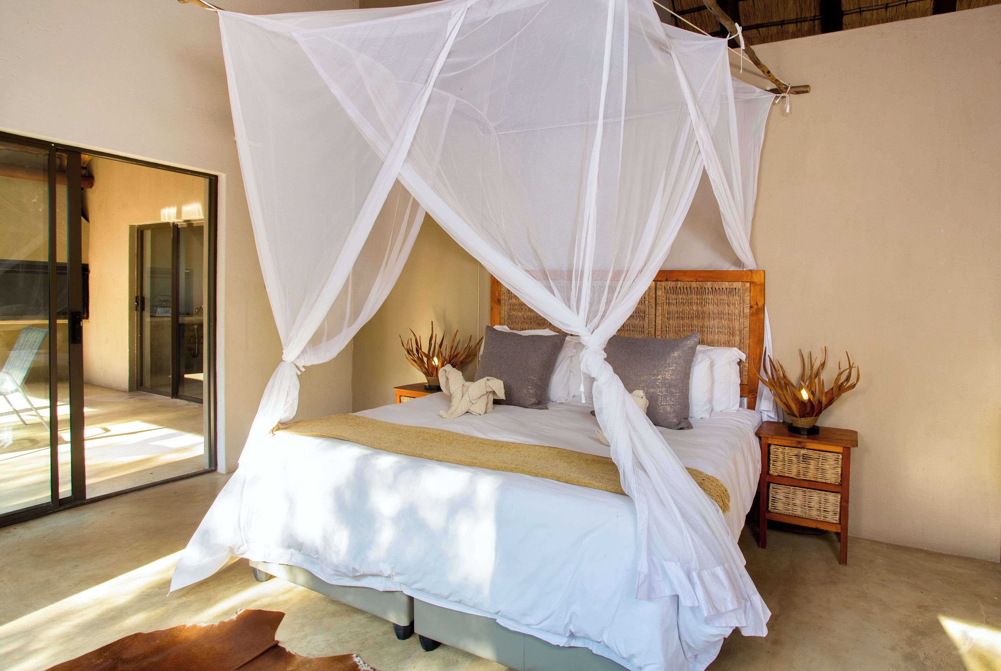 A bedroom with a bed under a white canopy, a wooden nightstand with a woven basket, and a view of a glass door leading outside.