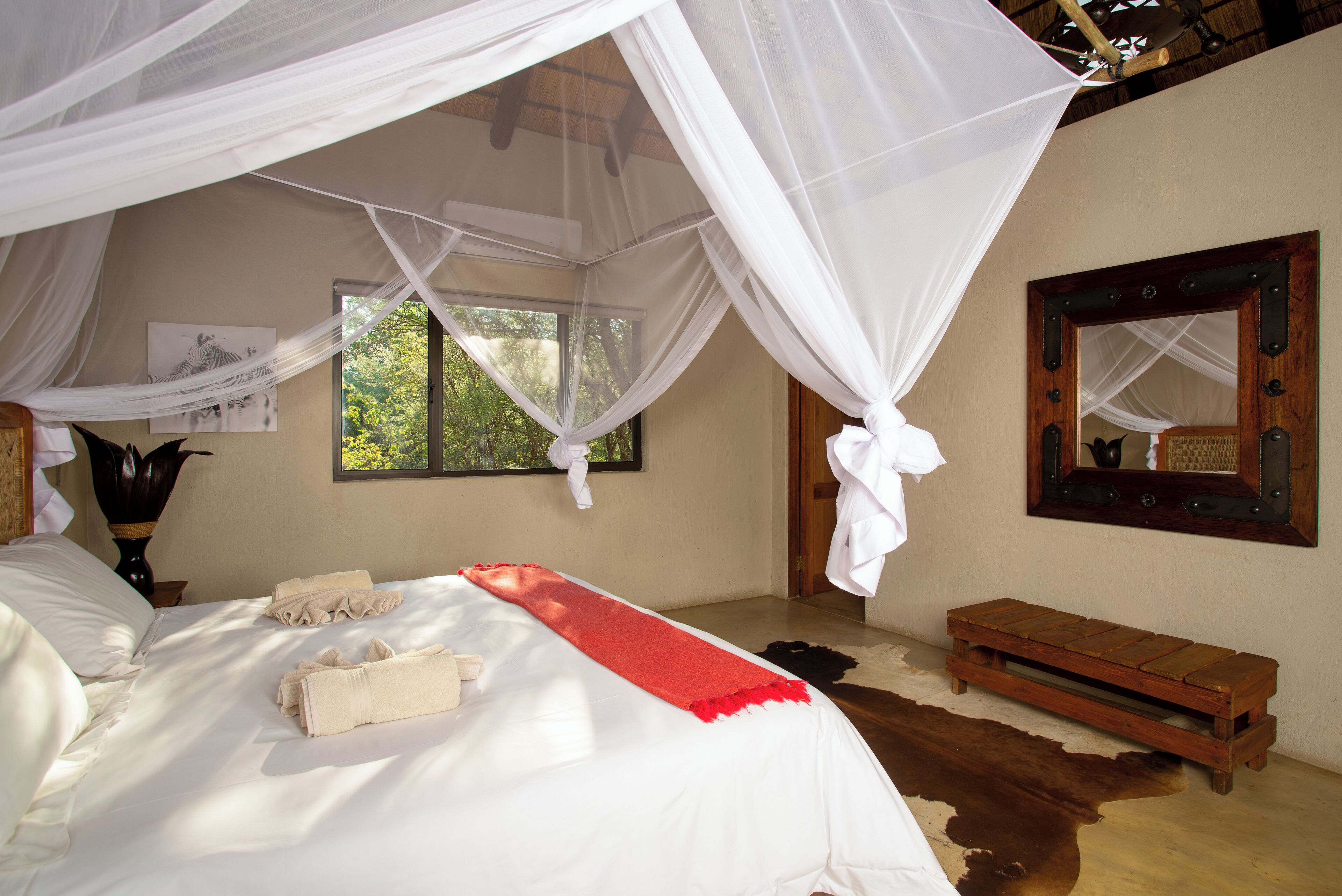 A bedroom with a canopy bed, a wooden bench, a mirror, and a window with a view of greenery.