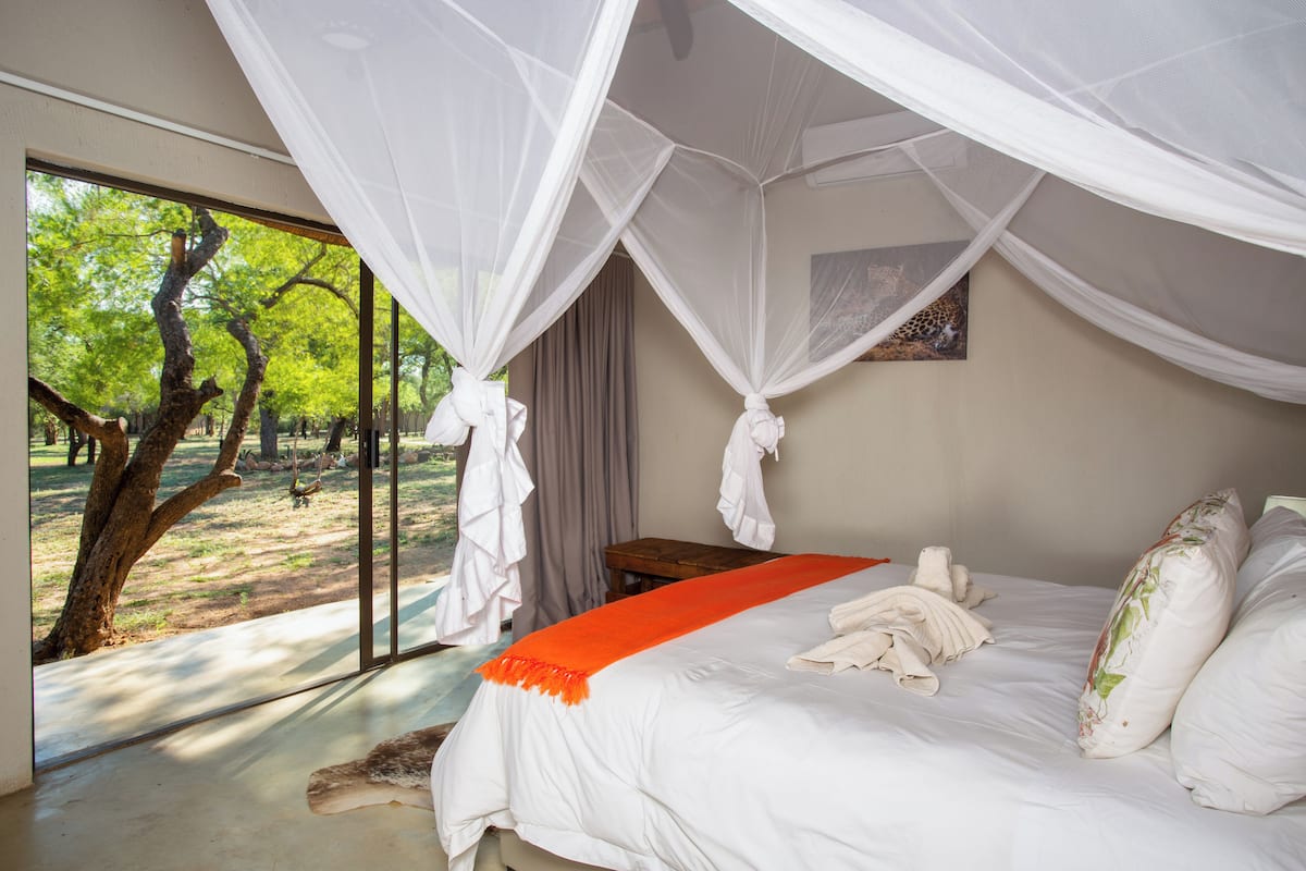 A bedroom with a large window, a bed with white bedding, a wooden nightstand, and a mosquito net.