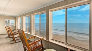 Ein Zimmer mit großen Fenstern und Blick auf einen Strand, Holzschaukelstühle und ein kleiner runder Tisch.