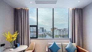 Quarto de hotel moderno com sofá, uma pequena mesa, um vaso de flores amarelas e vista para a cidade através de grandes janelas.