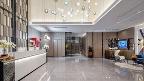 Um lobby moderno de hotel com balcão de recepção, área de estar com televisão e luminária decorativa no teto.