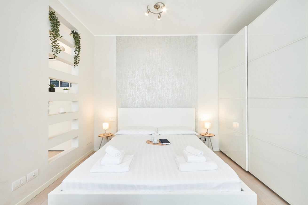A modern hotel room with a large bed, bedside tables, a wall-mounted shelf with plants, and a textured wall behind the bed.