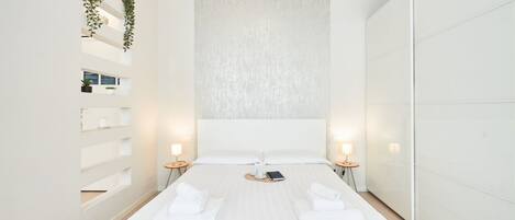 A modern hotel room with a large bed, bedside tables, a wall-mounted shelf with plants, and a textured wall behind the bed.