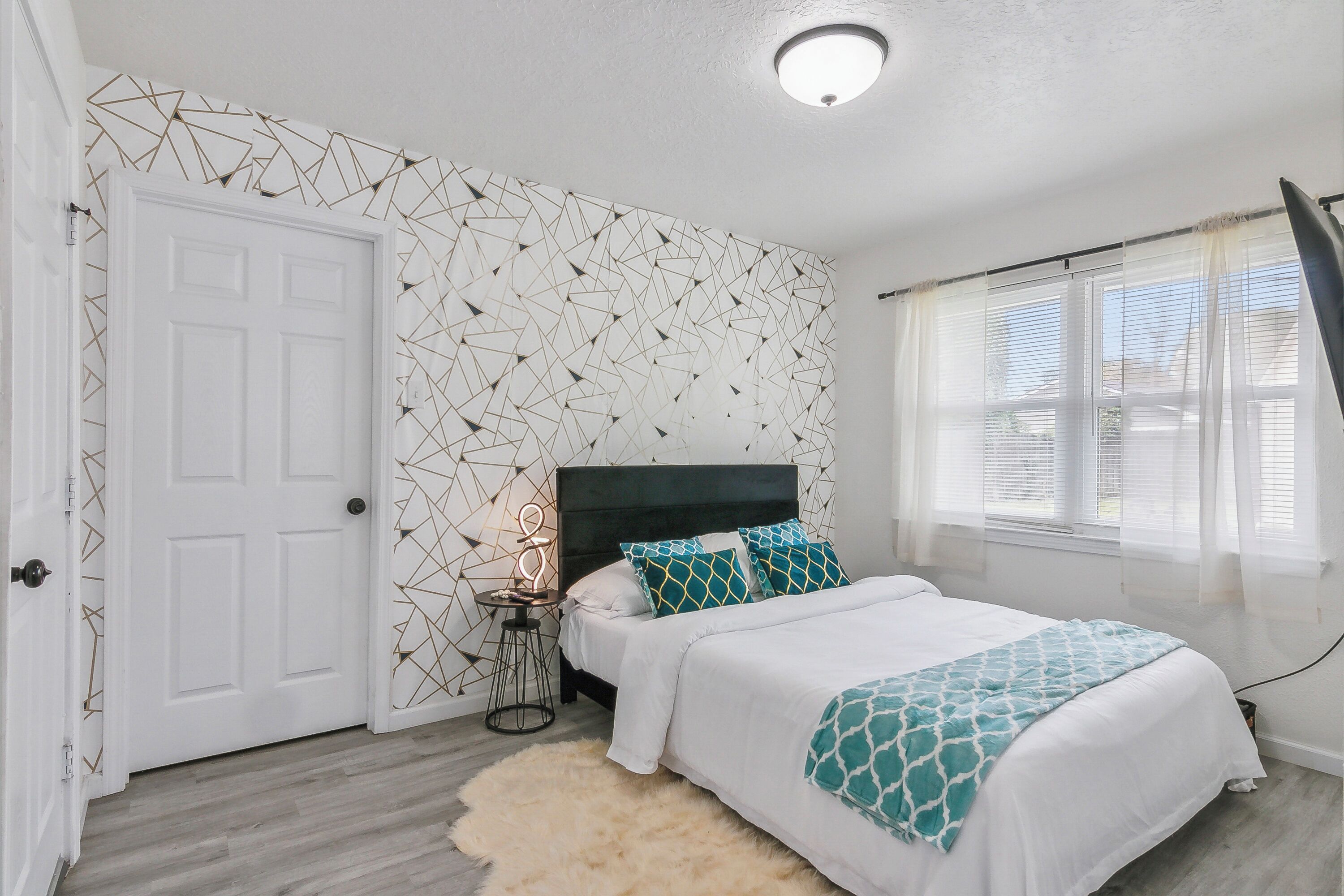A bedroom with a bed, a nightstand, a wall with a geometric pattern, a white door, and a window with blinds.