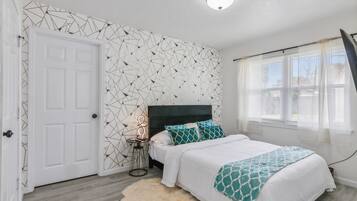 A bedroom with a bed, a nightstand, a wall with a geometric pattern, a white door, and a window with blinds.