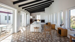 A spacious living area with a white sofa, wooden chairs, and a coffee table. Large windows provide ample natural light and views of the outdoors.