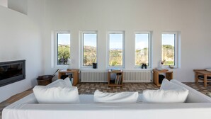 A modern living room with a white sofa, a fireplace, and a view of the countryside through large windows.