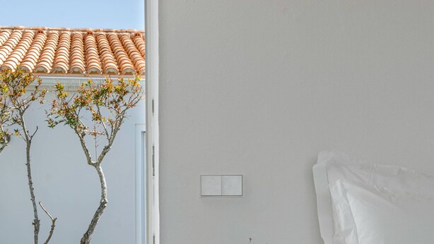 A bedroom with a bed, a bedside table with a vase of flowers, a view of a swimming pool, and a tree outside.