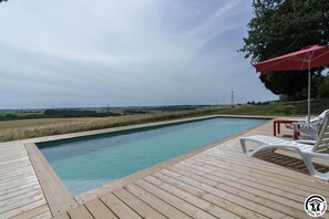 Een houten terras met een zwembad, witte ligstoelen en een rode parasol.